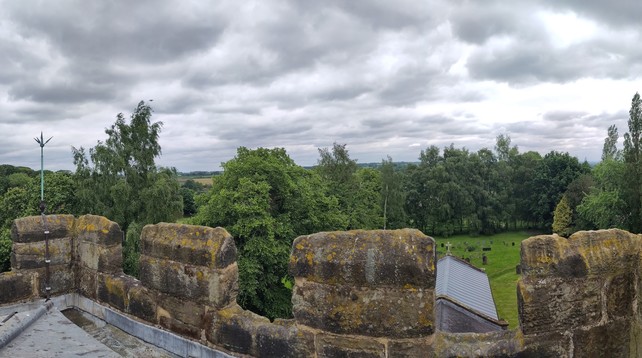 The first half of a panoramic picture looking east from the tower. You can see the parapets around the edge of the roof.

On the left there is a small lightning conductor rising from the parapet.

Slight to the right of the picture you can see the chancel roof with a cross on the far end and the churchyard beyond.

There are tall trees all around in full leaf, behind which you can see a few fields and hills.